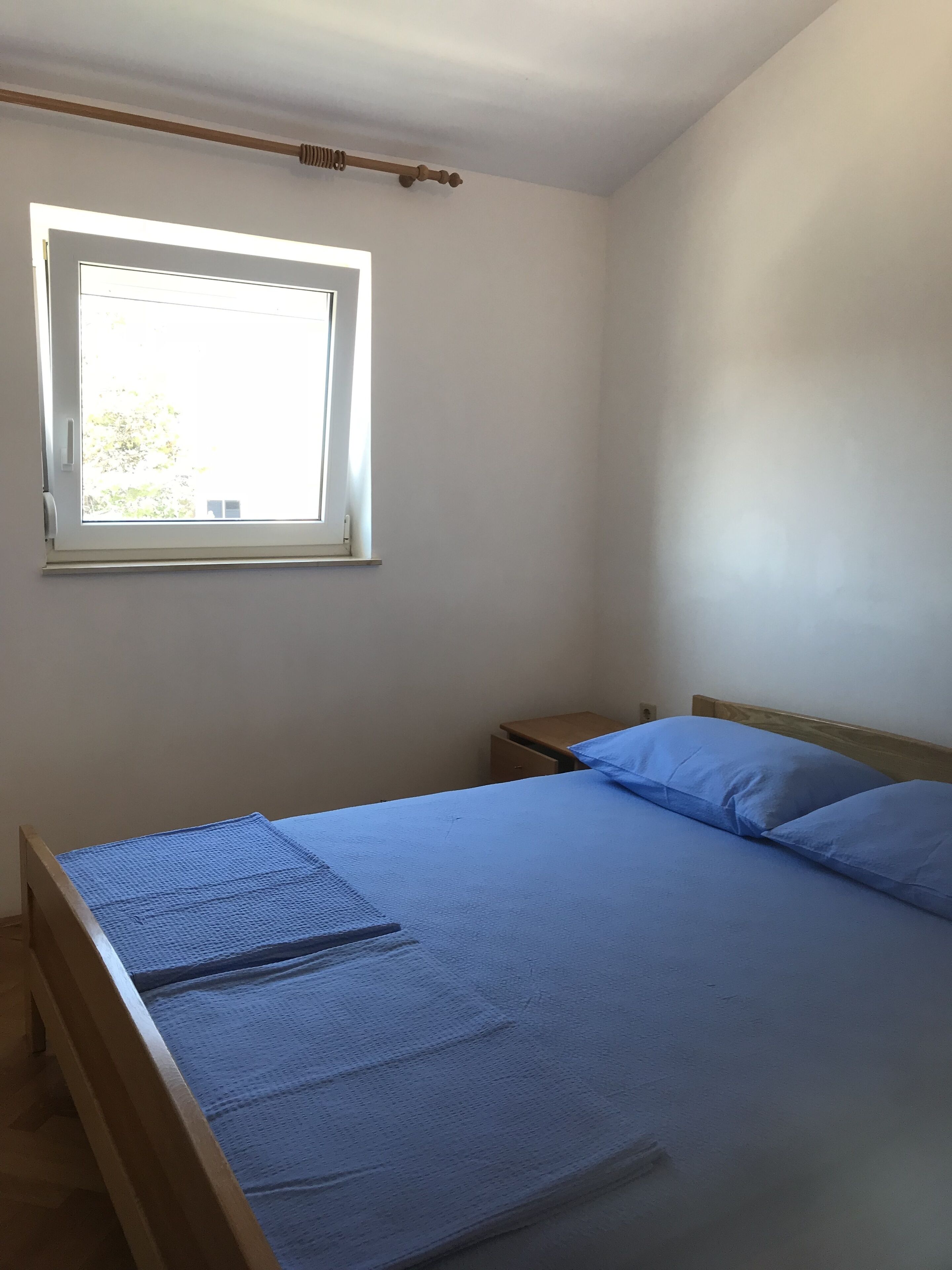 Ein Schlafzimmer mit einem Einzelbett, einem Holzkopfteil, einem Nachttisch, einem Fenster mit einer Holzjalousie und einer weißen Wand.