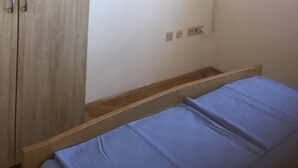 A bedroom with a wooden wardrobe, a small shelf, an air conditioning unit, and a bed with a blue mattress.