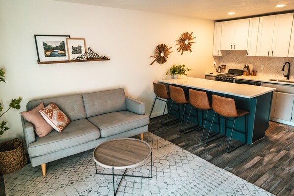 A modern kitchen with white cabinets, a central island with bar stools, a grey sofa, and a round wooden coffee table.