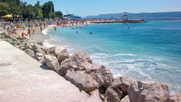 Ein Strand mit felsigem Ufer, klarem blauen Wasser und Menschen, die schwimmen und sonnenbaden.