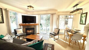 Una sala de estar moderna con chimenea, un televisor de pantalla plana, un comedor con mesa y sillas, y ventanas amplias que ofrecen vistas al exterior.