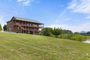 A two-story wooden house with a balcony, a garage, and a grassy hill with trees and a lake in the background. - Wonderland Cove by Jackson Mountain Rentals (Dandridge)