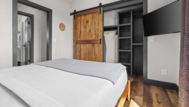 A bedroom with a bed, a wardrobe with a wooden door, a TV mounted on the wall, and a bathroom visible through an open door. - 2 Luxe Nashville Homes | Near to Broadway (Nashville)