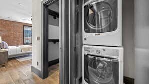 A laundry room with two stacked washing machines, a bed with a view to a brick wall, and a door leading to another room. - 2 Luxe Nashville Homes | Near to Broadway (Nashville)