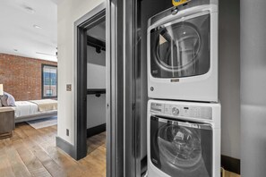 A laundry room with two stacked washing machines, a bed with a view to a brick wall, and a door leading to another room. - 2 Luxe Nashville Homes | Near to Broadway (Nashville)