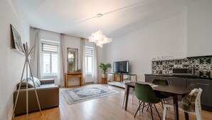 A modern living room with a sofa, dining table, chairs, a TV, and a kitchen area with a microwave. - Baross Boutique Apartman Urban Rooms (Gyor)