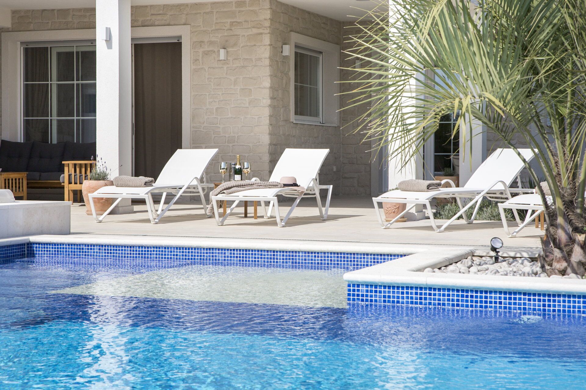 A pool area with white lounge chairs, a stone wall, and a palm tree.