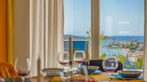 Mesa de comedor con copas de vino y platos, con vistas a una ciudad costera con edificios y barcos.