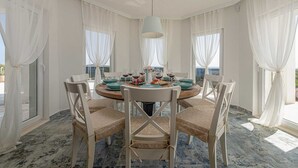 A round dining table with white chairs, set for four people, in a room with large windows and white curtains.