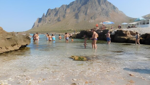 A beach with clear shallow waters, rocky formations, and a mountain in the background.