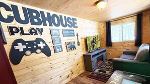 Interior acollidor d'una cabana amb una paret de fusta que mostra els cartells 'CUBHOUSE PLAY' i 'GAME ON', una llar de foc, un sofà i una televisió.
