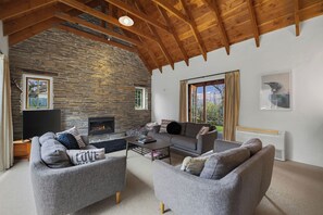 A living room with a stone wall, wooden beams, a fireplace, and a large window. - Maytime Cottage - Arrowtown Holiday Home (Arrowtown)