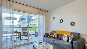 Ein Wohnzimmer mit einer Couch, einem Glastisch und einer Glastür, die auf einen Balkon mit Stühlen und einer Pflanze führt.