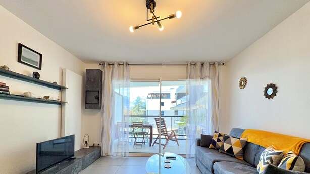 Ein Wohnzimmer mit einer Couch, einem Glastisch, einem Fernseher und einem Balkon mit Tisch und Stühlen.