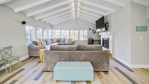 Un salon spacieux doté d’un haut plafond, de grandes fenêtres et d’un coin salon confortable avec un canapé d’angle et un pouf.