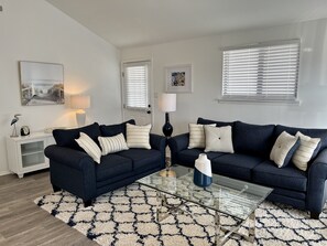 A living room with two dark blue sofas, a glass coffee table, a patterned rug, a white cabinet, and a door with blinds. - Renovated Home with Heated Pool 3 Bed/2 Bath - Sleeps 6 (Avalon)