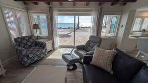 Un salon comprenant un canapé, un fauteuil et un pouf, un ventilateur de plafond et une vue sur la plage grâce à de grandes fenêtres.