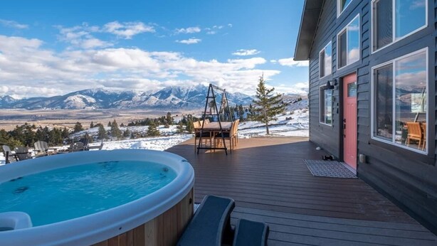 A hot tub on a wooden deck with a mountain view, a dining area, and a building with large windows. - Sky Cabin • Mountain Sauna • Hot Tub • Yellowstone (Emigrant)
