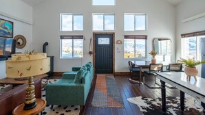 A spacious living room with a high ceiling, large windows, a dark door, a teal sofa, a wooden coffee table, and a dining area with a long table and chairs. - Sky Cabin • Mountain Sauna • Hot Tub • Yellowstone (Emigrant)
