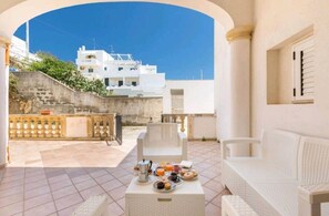 A terrace with a white sofa, a table with breakfast items, and a view of white buildings under a clear sky.