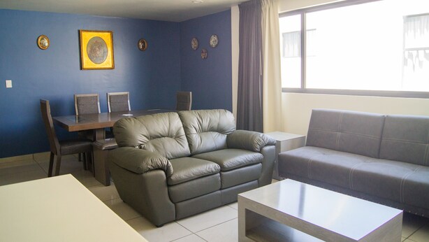 A living room with a sofa, a coffee table, a dining table with chairs, and a large window with curtains. - Villas Analco Apartments (Puebla)