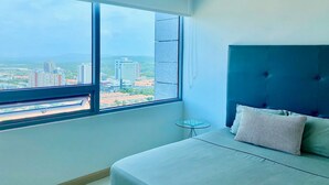 A bedroom with a large window, a bed, a small table, and a city view. - Apartamento Vacacional en Barranquilla (Barranquilla)
