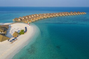 An overwater resort with thatched-roof bungalows connected by walkways, a sandy beach, and clear turquoise waters.