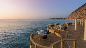A wooden deck with tables and chairs overlooking the sea, with a thatched-roof structure in the background. - Emerald Faarufushi Resort & Spa (Faarufushi)