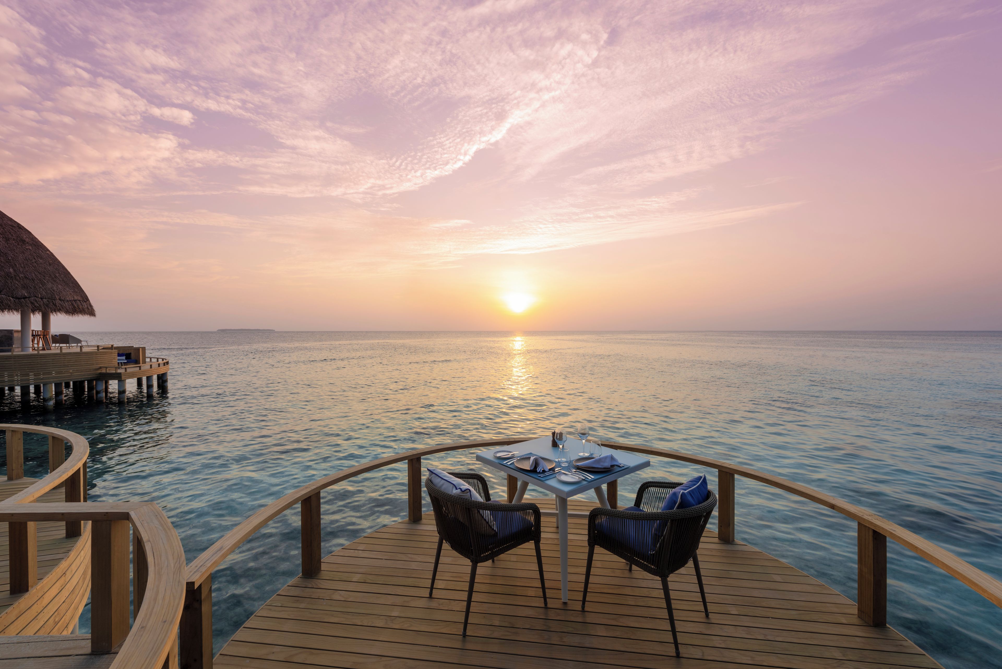 A wooden deck with a table and chairs overlooking a calm sea at sunset.