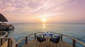 A wooden deck with a table and chairs overlooking a calm sea at sunset.