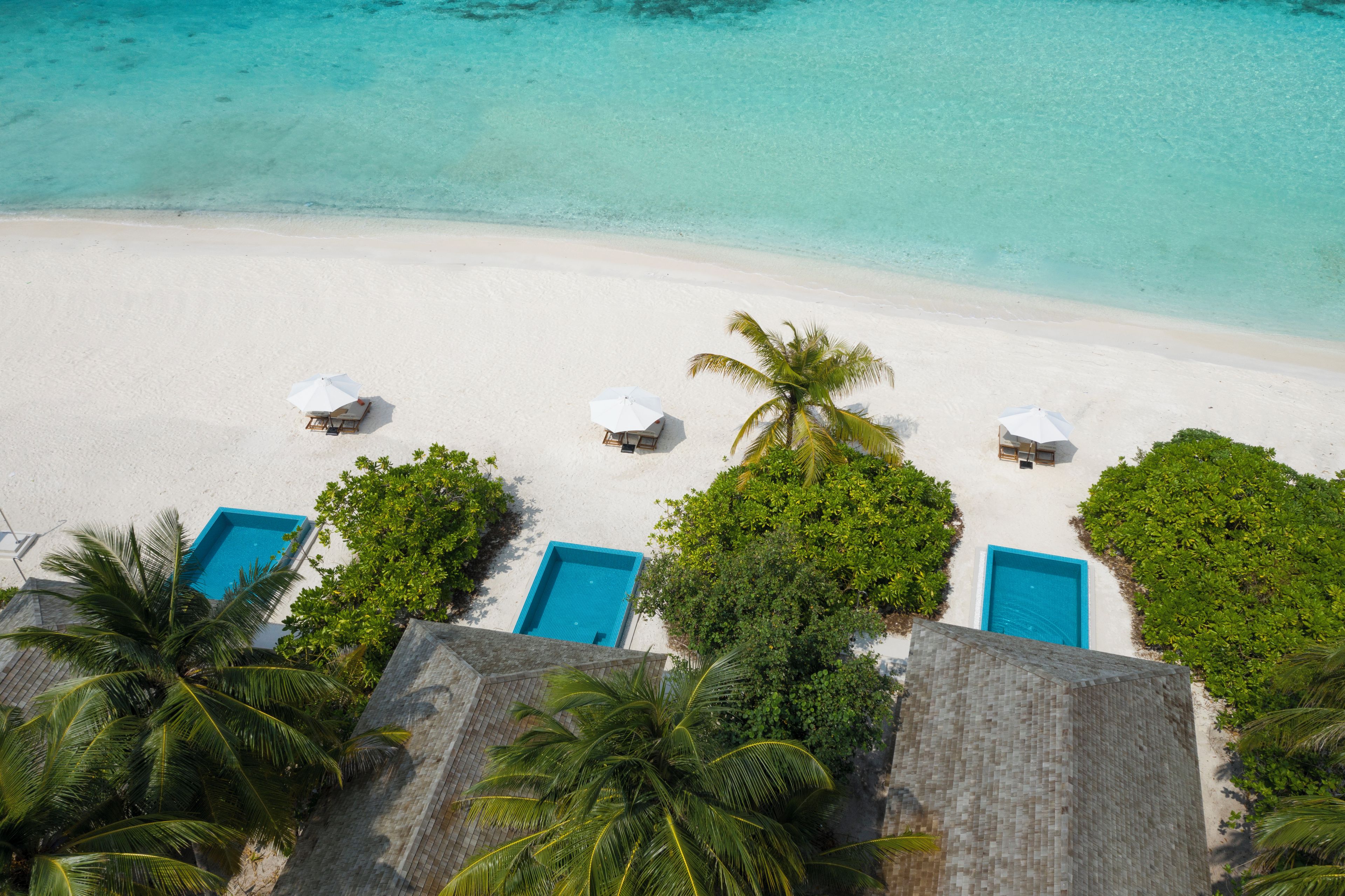 An aerial view of a beach with white sand, clear turquoise water, and several blue pool areas.