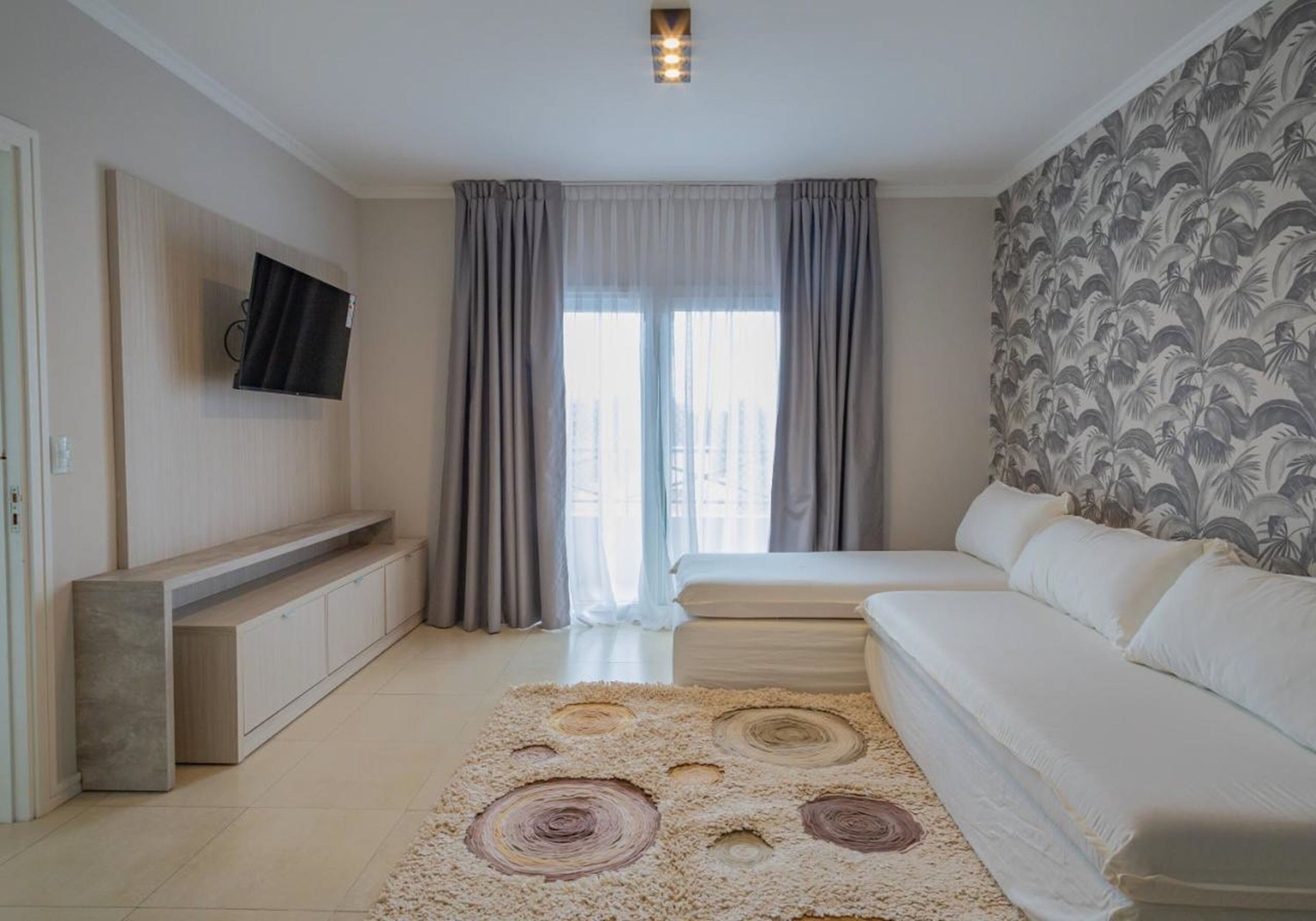 Un dormitorio moderno con cama, sofá, televisor montado en la pared y un amplio ventanal con cortinas.