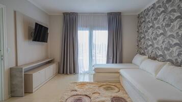 Un dormitorio moderno con cama, sofá, televisor montado en la pared y un amplio ventanal con cortinas.