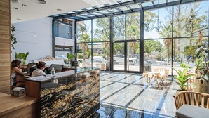 A modern hotel lobby with a marble reception desk, wooden furniture, and large windows offering a view of the street and greenery. - Garden Suites Pinamar (Pinamar)