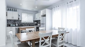 A modern kitchen with white cabinets, a wooden dining table, and a large window with sheer curtains. - Nautilus on the Inlet (Inverloch)