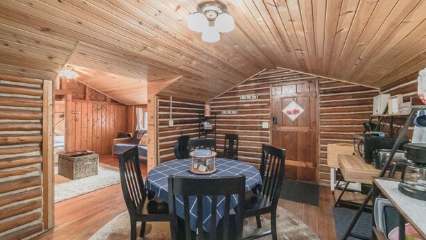 Interior acogedor de una cabaña de troncos, con comedor que incluye una mesa redonda y sillas, techo de madera y una pequeña cocina con electrodomésticos básicos.