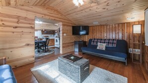 Un acogedor interior de cabaña con techo de madera, un comedor, una sala de estar con sofá y televisor, y una pequeña cocina.