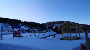 A snow-covered playground with swings and a slide, surrounded by houses and a forested hill.