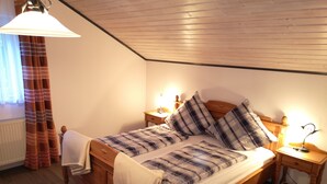 A wooden bed with a plaid bedspread and pillows, flanked by bedside tables with lamps, in a room with wooden flooring and a sloped ceiling.