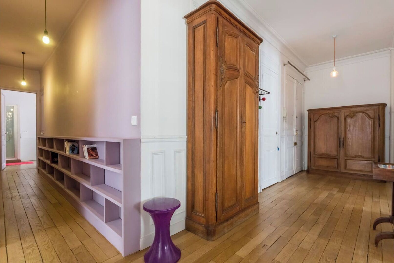 A spacious room with wooden flooring, a tall wooden wardrobe, and a purple side table.