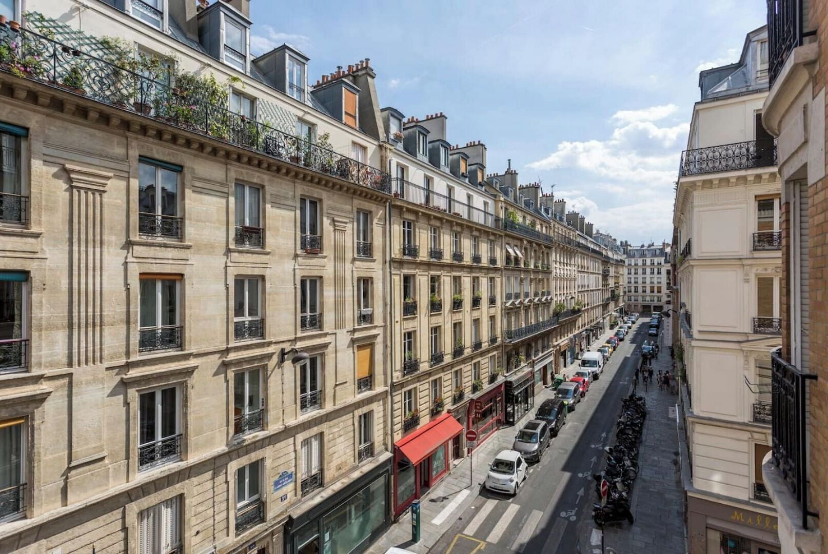 Une rangée de maisons de ville européennes classiques, toutes de style architectural uniforme, avec des balcons ornés de plantes, une rue bordée de voitures garées et un ciel dégagé.