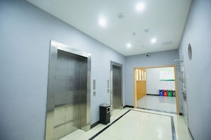 Un pasillo con dos ascensores, un cesto de basura y una puerta que da a otra habitación con un tablón de anuncios.