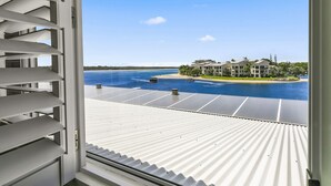 Une vue depuis une fenêtre avec des stores blancs, donnant sur une étendue d’eau où l’on aperçoit des bateaux, une plage de sable et une rangée de bâtiments bordée d’une pelouse verte.
