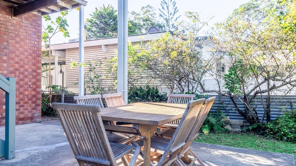 Un patio al aire libre con mesa y sillas de madera, rodeado de follaje y un muro de ladrillo.