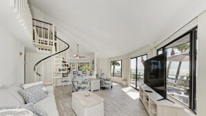 A spacious living area with a staircase, a large TV, and a balcony with a view of the sea. - The Turtle House-Miles and Miles of Private Beach-Sunset Views! (Englewood)