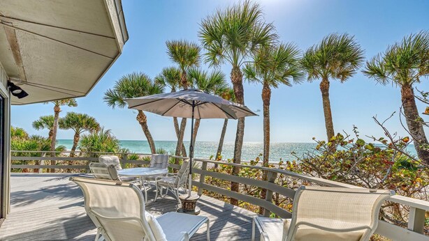 A deck with outdoor seating, a table, and an umbrella, overlooking a beach with palm trees and the ocean. - The Turtle House-Miles and Miles of Private Beach-Sunset Views! (Englewood)