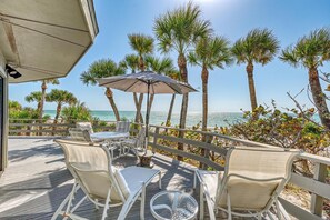 Une terrasse avec des sièges, une table et un parasol, offrant une vue sur une plage bordée de palmiers et sur l’océan.