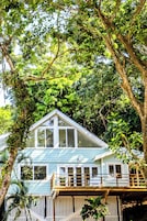 Una casa azul claro con balcón blanco, rodeada de abundante follaje y árboles verdes.