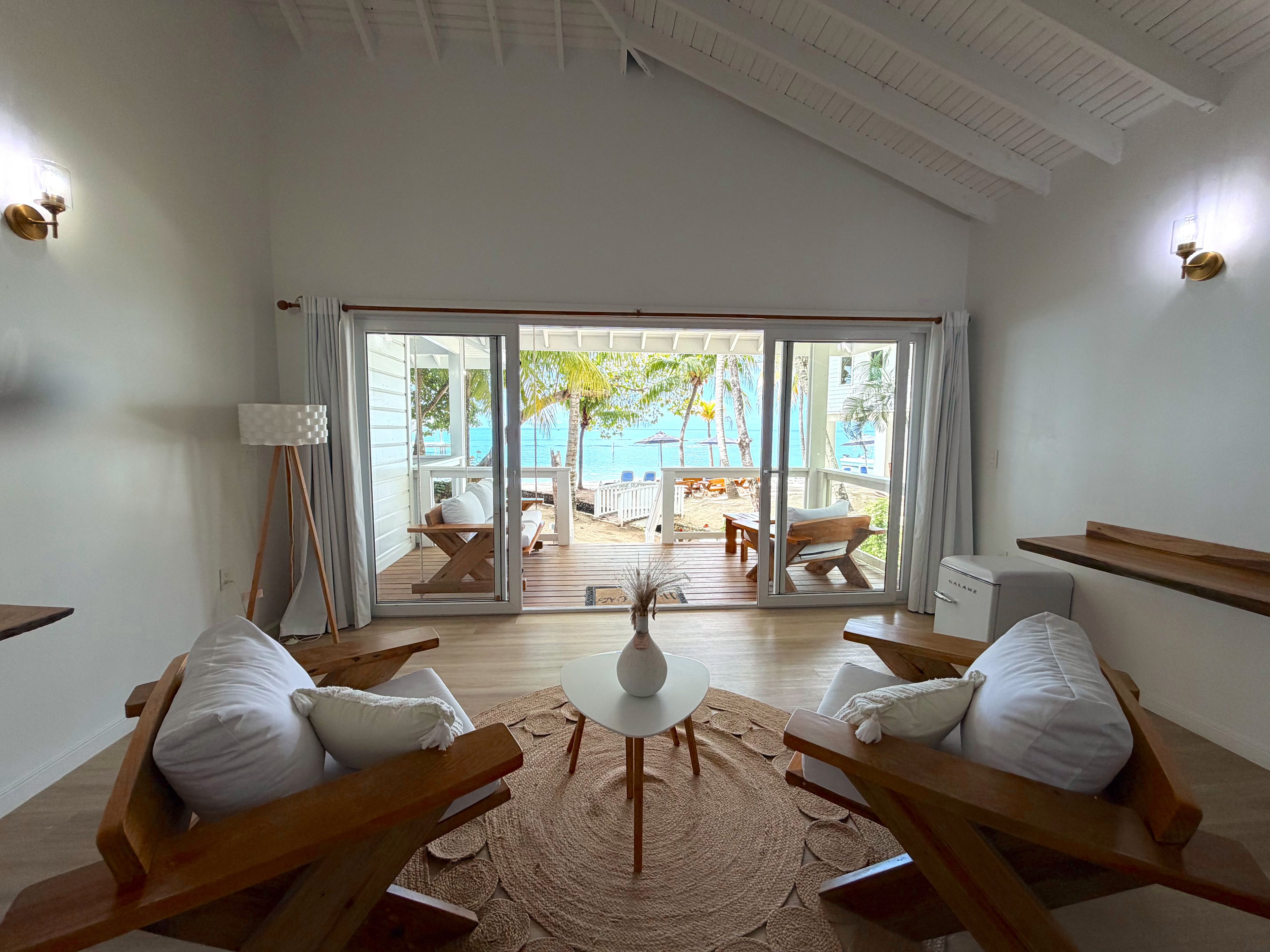 A room with a view of a beach, a wooden table, two chairs, and a lamp.