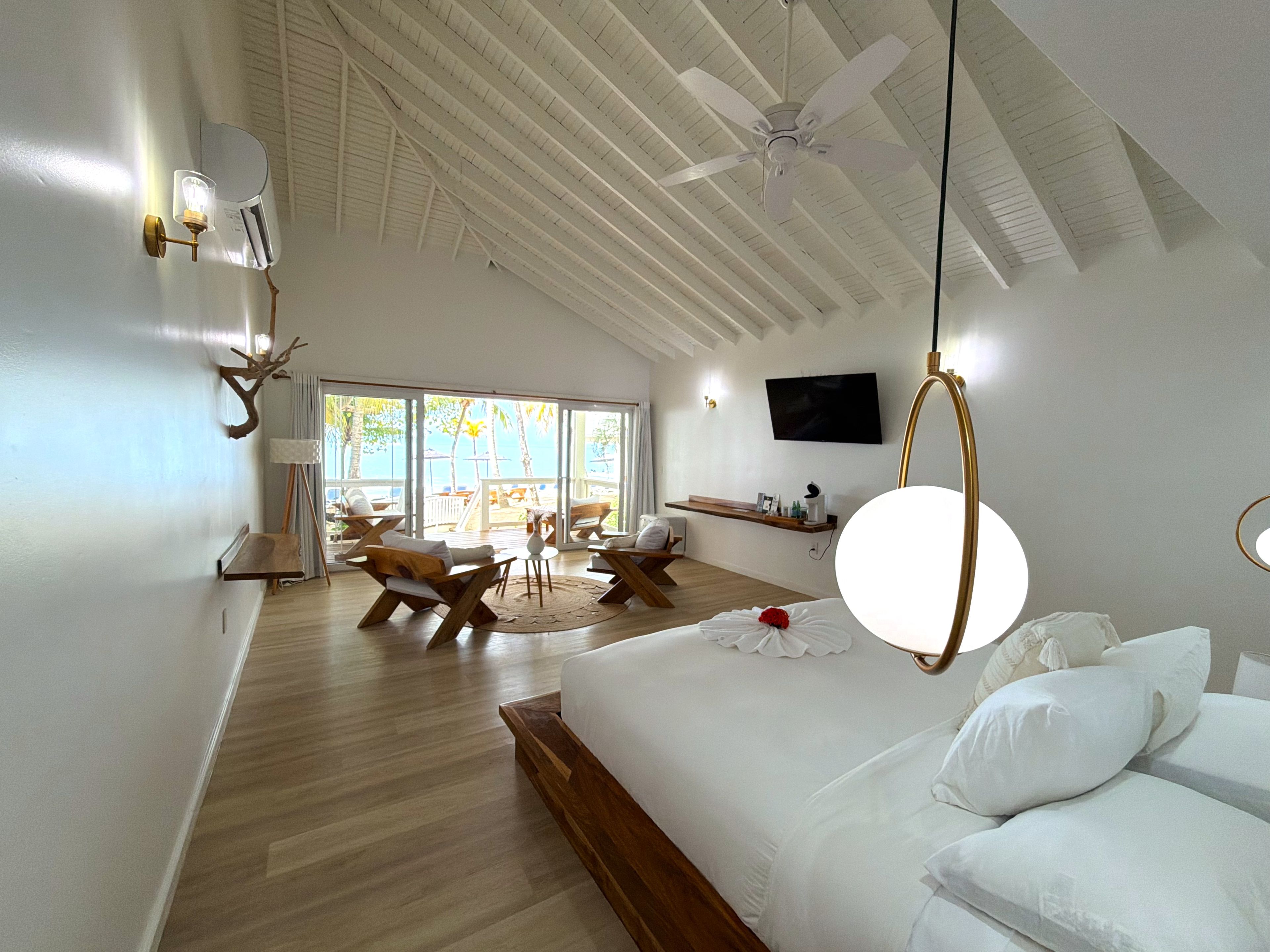 A spacious room with a large bed, a ceiling fan, a TV, and a view of the beach.